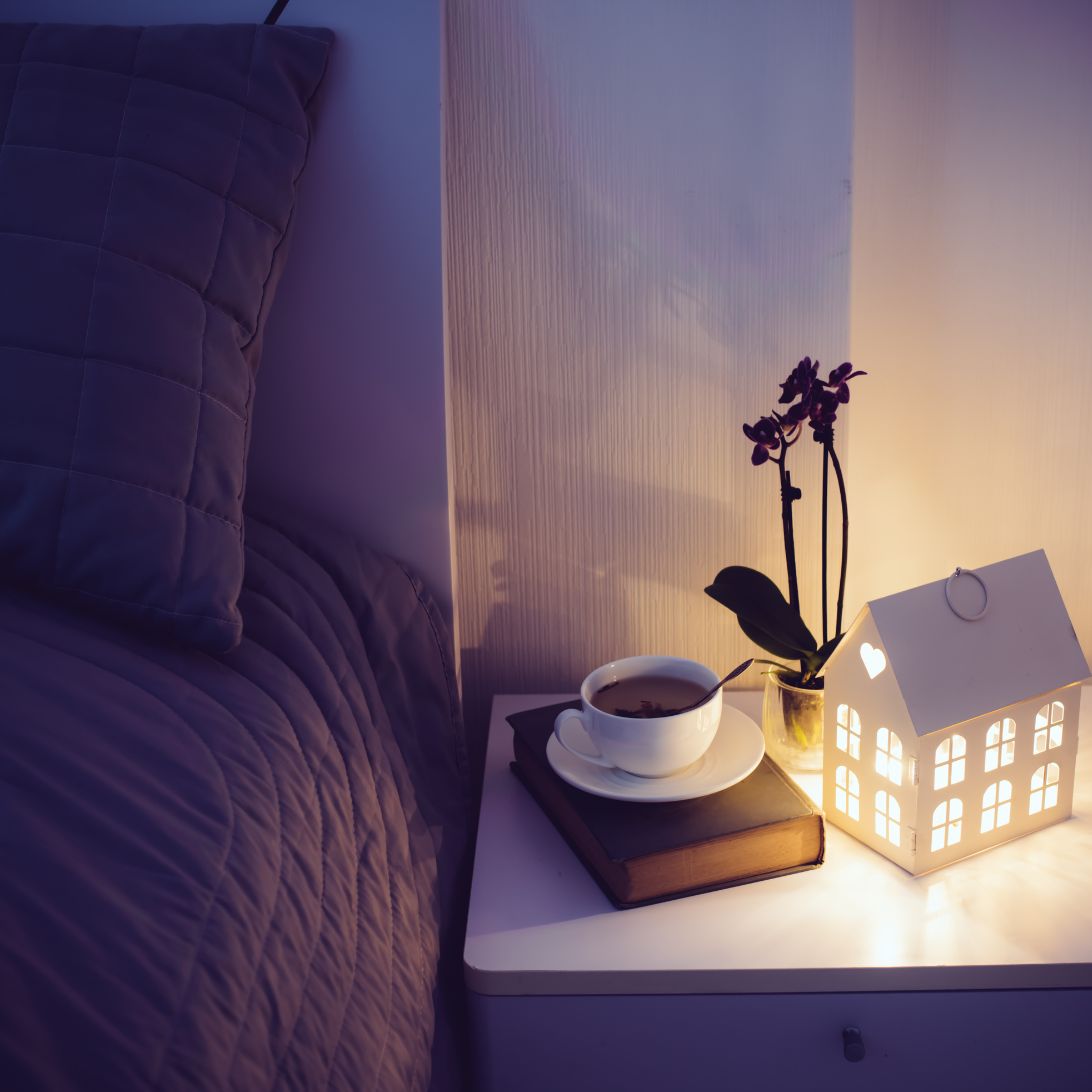 Bed met nachtkastje met kopje koffie, lamp in de vorm van een huisje en plantje in kleine slaapkamer