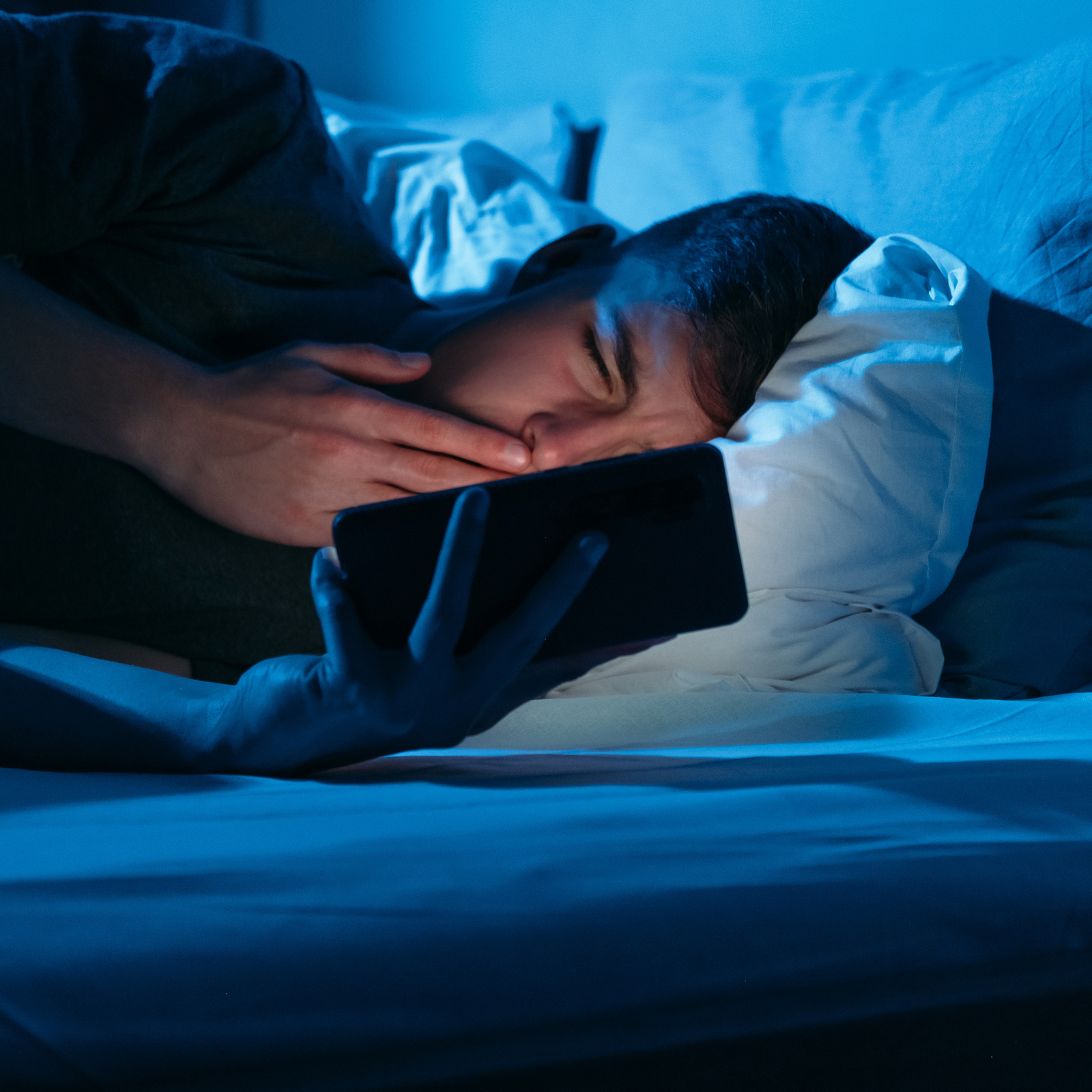 Gapende man gebruikt zijn telefoon in bed, blauw ligt schijnt op zijn gezicht, bewijst de invloed van technologie op slaap